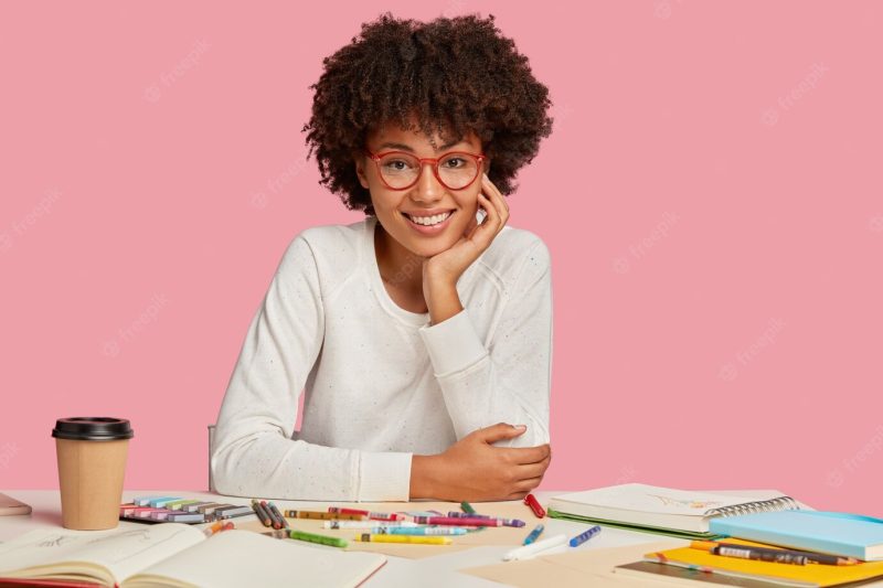 Image of pleasant looking ethnic cheerful woman with afro hairstyle, keeps hand on cheek, wears spectacles and white clothes, makes sketches in notepad, models at desktop over pink wall Free Photo