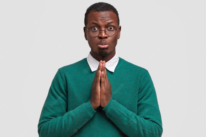 Image of sad gloomy African American man keeps palms pressed together, asks for help in close friend, purses lower lip, has dark healthy skin Free Photo