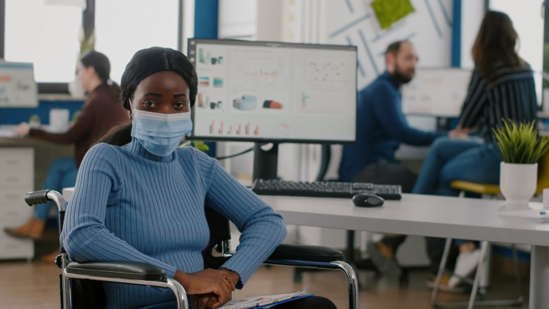 Immobilized paralyzed handicapped African employee looking at camera wearing protection mask during … Free Photo