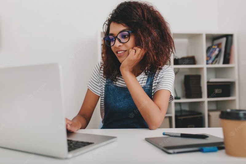 Indoor portrait of black young woman in glasses surfing in internet sitting in front of shelves with documents Free Photo