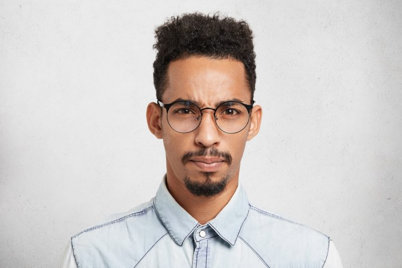 Indoor portrait of displeased discontent upset male model with trendy hairdo, mustache, beard, wears round glasses Free Photo