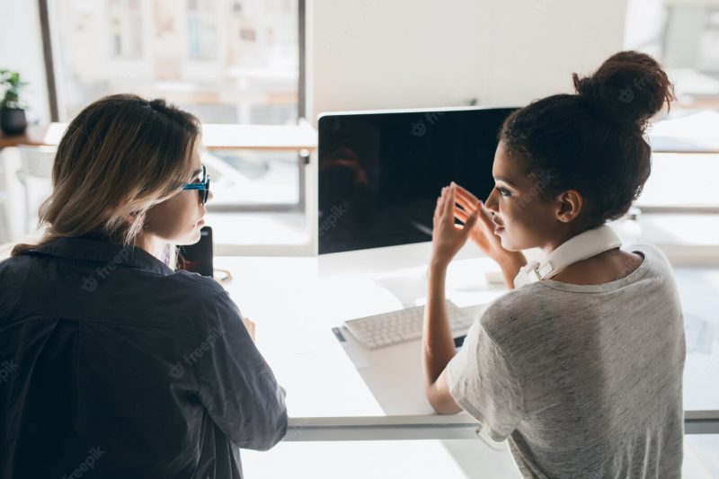 Indoor portrait from back of two women sitting in front of computer with black screen. african female office worker in headphones talking with colleague beside window. Free Photo