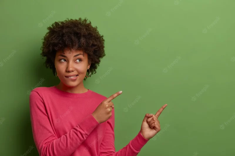 Indoor shot of curious african american woman bites lips, sees something good, indicates fingers at blank space on green wall, shows place for your product, company banner or promotion Free Photo