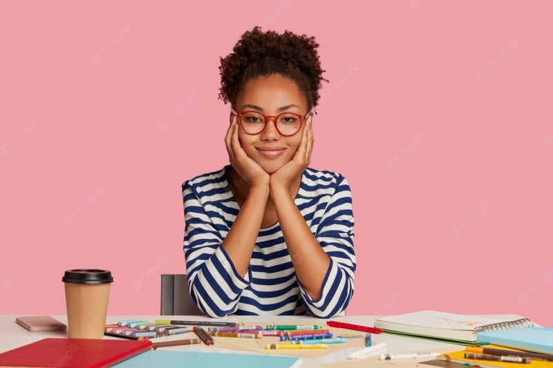 Indoor shot of dark skinned fashion designer with crisp hair, touches cheeks with both hands, wears striped clothes, works on new creative project, poses at workplace against pink wall Free Photo