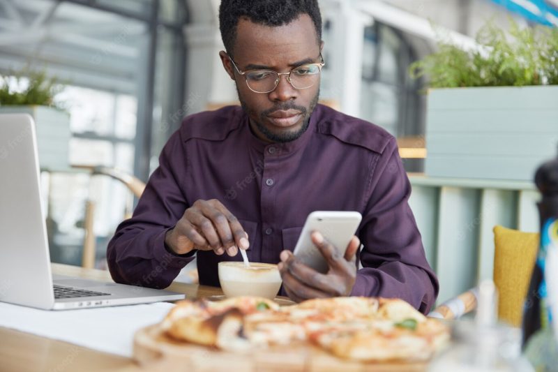 Indoor shot of dark skinned serious young african male enterpreneur focused into screen of mobile phone, drinks latte, reads attentively news on internet website Free Photo