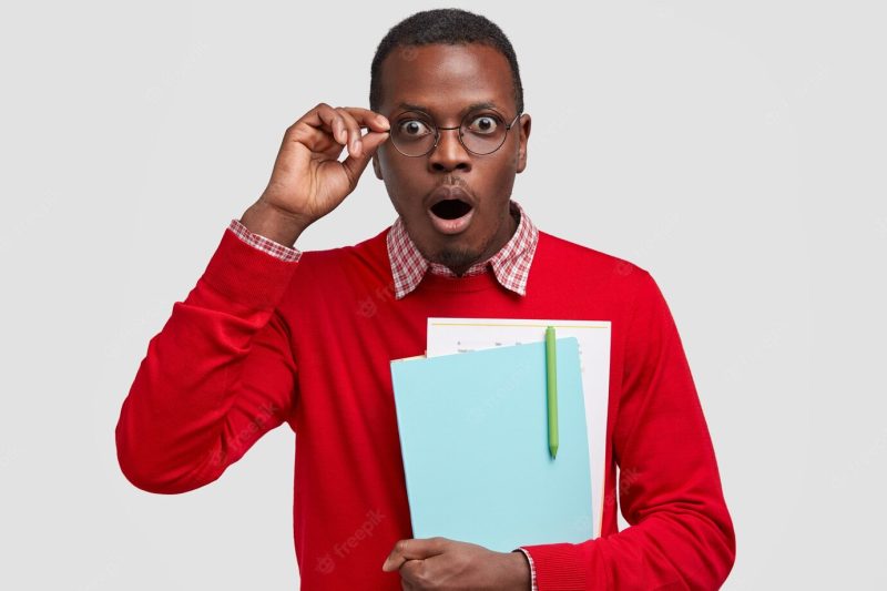 Indoor shot of stupefied dark skinned guy keeps jaw dropped, wears red clothes and spectacles, carries textbooks, writes scientific report Free Photo