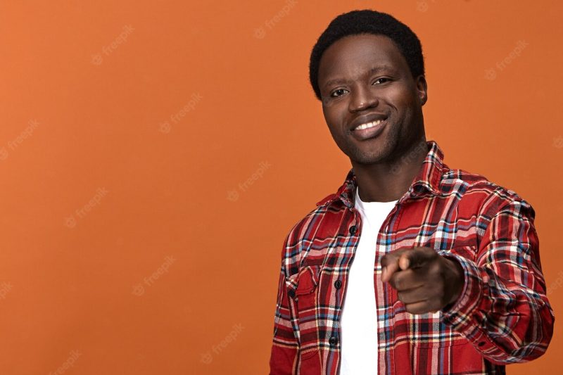 Isolated handsome cheerful young dark skinned black male wearing stylish plaid shirt smiling broadly and pointing his fore finger as if choosing you. selective focus Free Photo