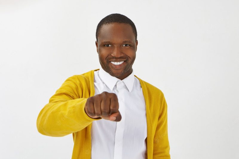 Isolated shot of happy positive young afro american man wearing stylish clothes posing , smiling broadly and holding clenched fist in front of him, ready to bump knuckles while greeting you Free Photo