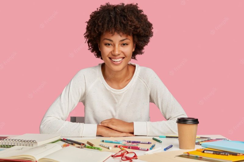 Isolated shot of hilarious dark skinned woman has afro haircut, dressed in casual white jumper, sits at desktop with creative chaos, drinks takeaway coffee, uses crayons, has toothy smile on face Free Photo