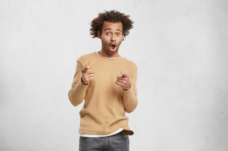 Joyful mixed race man wears sweater, points finger at camera, chooses you, opens mouth widely Free Photo