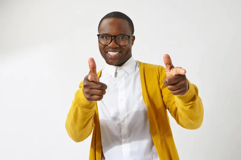 Joyful young afro american male hipster wearing stylish eyeglasses and cardigan smiling broadly, pointing index fingers , having positive friendly facial expression Free Photo