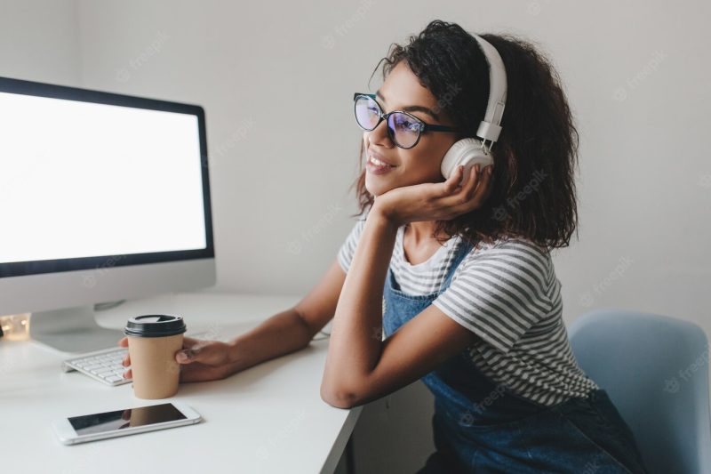 Lovely mulatto girl sitting at the table with computer and smartphone on it and propping up face with hand Free Photo