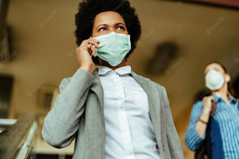 Low angle view of african american businesswoman walking through public corridor with protective mask and communicating over mobile phone Free Photo