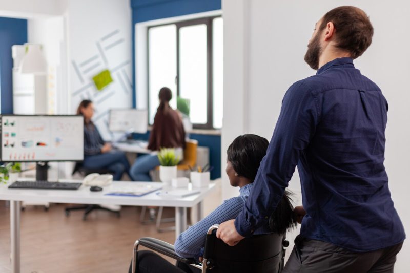 Man employee helping his invalid african colleague coming in workplace pushing wheelchair near desk Free Photo