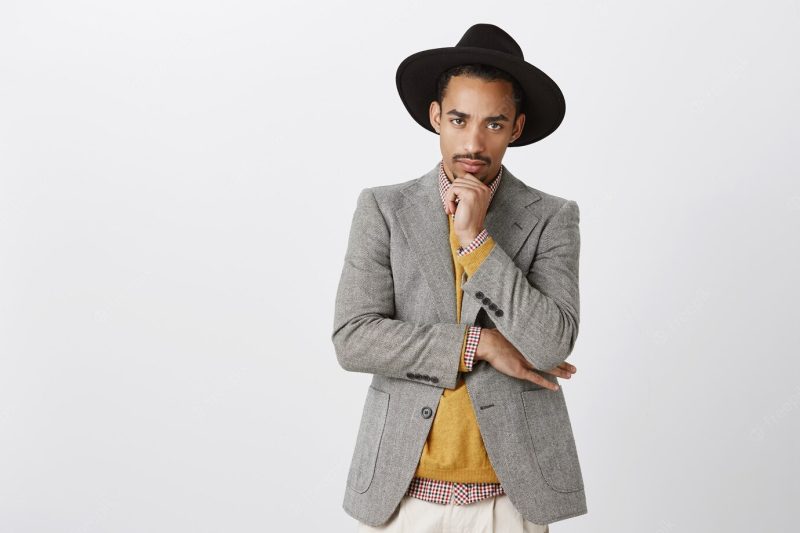 Man gets serious when it comes to job. portrait of good-looking focused young dark-skinned male in stylish hat and outfit, holding hand on chin, staring, making hard decision over gray wall Free Photo