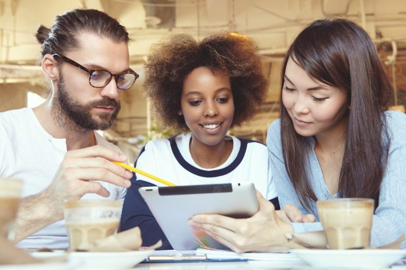 Man in glasses presenting business strategy on tablet pc, showing information on screen with pencil while his asian and african colleagues listening to him with attention, sitting at co-working space Free Photo