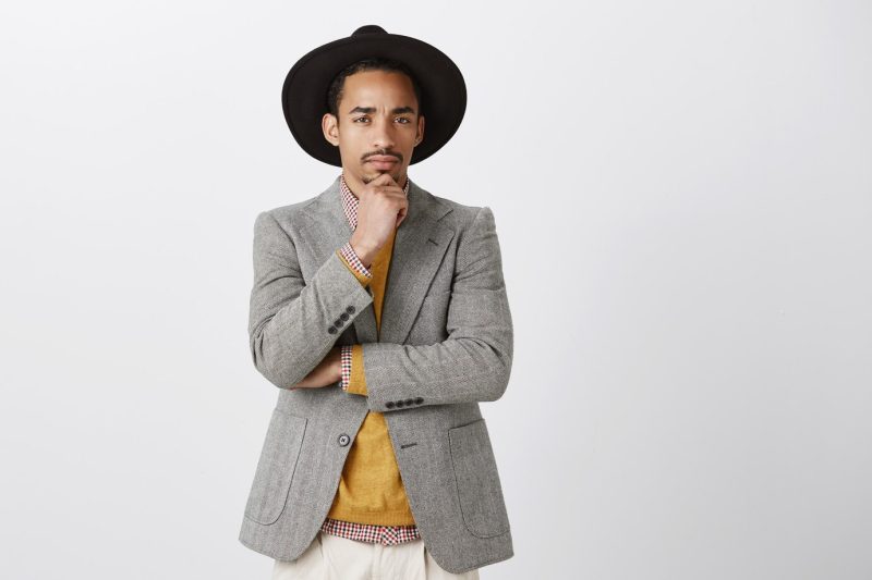 Man is not sure about you. portrait of doubtful good-looking male in trendy jacket and hat, holding hand on chin, frowning, looking suspicious while thinking, having doubts or disbelief over gray wall Free Photo