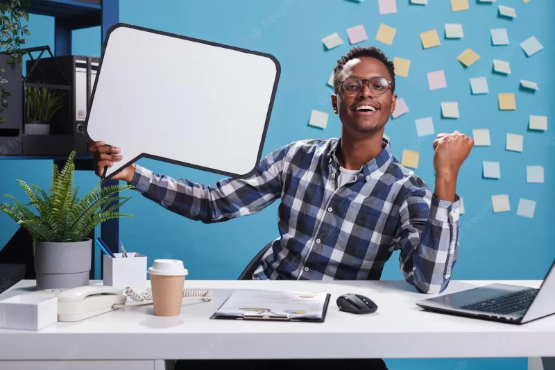 Marketing company happy smiling young adult person holding bubble sign while clenching fist triumphant.management department team leader with cardboard speech bubble at work. Free Photo