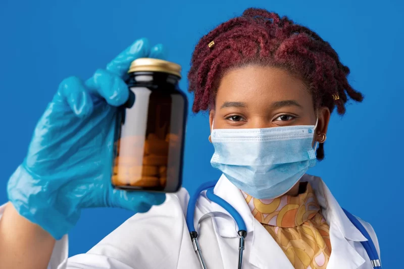 Masked african american woman doctor showing glass jar of medicine against blue background Free Photo