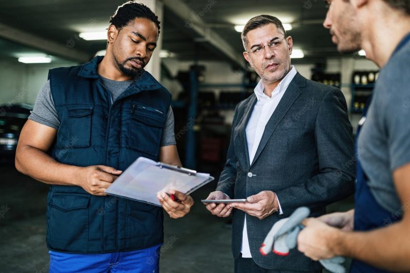 Mid adult businessman with touchpad communicating with car mechanics in auto repair shop Free Photo