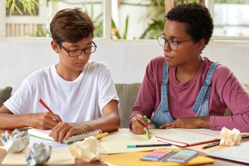 Mixed race classmate study together, write in notebook, rewrite information from papers, prepare for school exam, wear casual clothes, pose at desktop, spend time together. people, assistance concept Free Photo