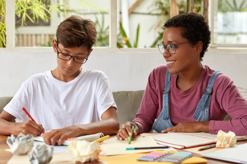 Multi ethnic  group mates write common essay, sit at table with papers and notepad, dressed in casual clothing. Asian boy receives consultancy from dark skinned friend, check information from documents Free Photo