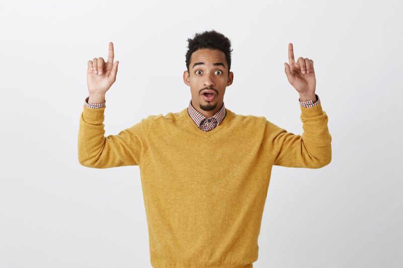 Never seen copy space more beautiful. portrait of attractive emotive African-American male student in yellow sweater raising index fingers, pointing upwards with dropped jaw and surprised expression Free Photo