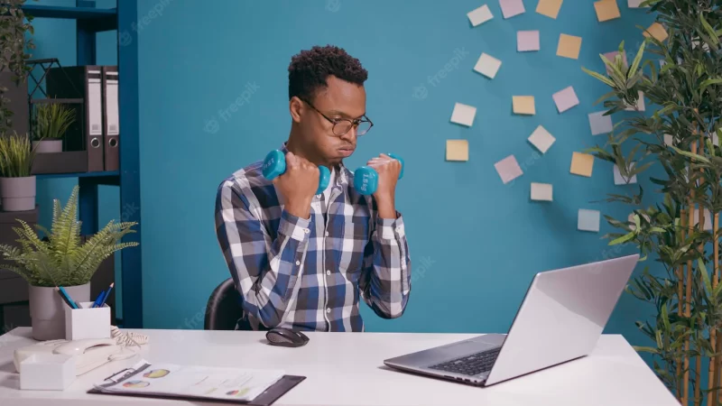 Office worker exercising with dumbbells to train muscles while he uses laptop at desk. employee lifting weights and doing physical activity with fitness exercise, working on computer for business. Free Photo