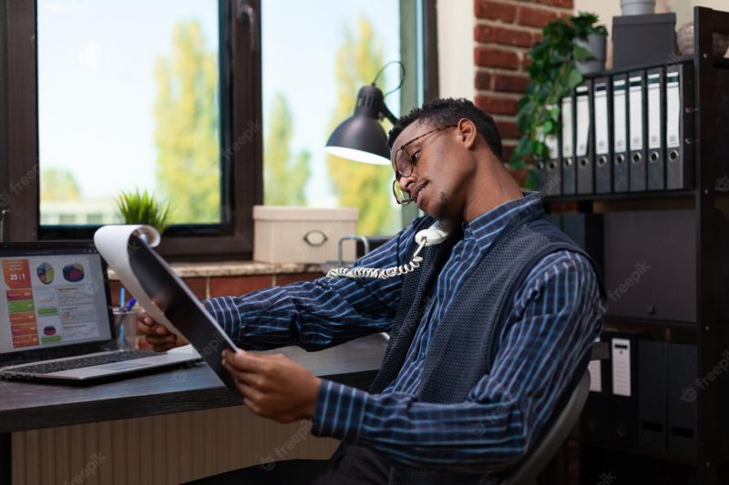 Office worker holding phone reciever on shoulder with tilted head looking at clipboard with business analytics. startup employee sitting at desk with laptop discussing profit with manager on the telephone Free Photo