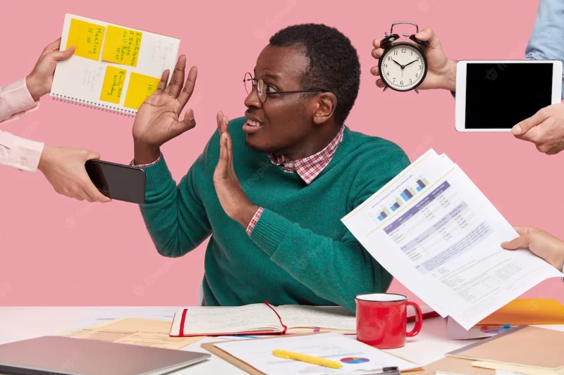 Oh no, not now! stressed dark skinned man has much work in office, asked by many people at one time who remind about preparing business project, hold alarm clock Free Photo