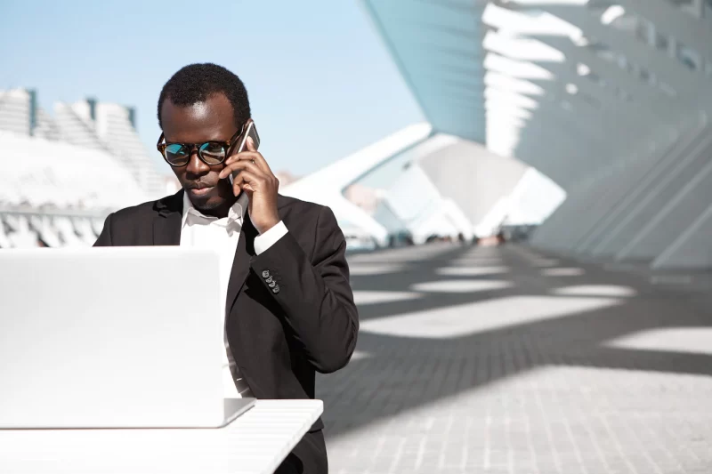 Outdoor portrait of successful confident young african entrepreneur or corporate worker in black suit and stylish shades having phone conversation and working remotely on laptop pc at urban cafe Free Photo