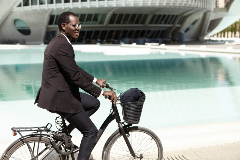 Outdoor summer portrait of handsome young black european office worker in sunglasses cycling on his bike to work in urban surroundings on, having fun, feeling carefree and relaxed expression Free Photo