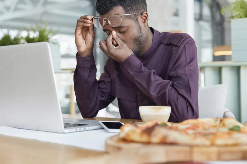 Overwork, tiredness concept. exhausted dark skinned male employee sits in front of laptop, works on new project for long time, has pain in eyes Free Photo