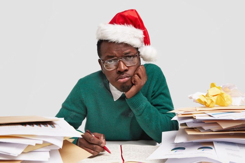 Overworked sad African American man keeps hand on cheek, wears Santa Claus hat, optical spectacles, writes down in diary Free Photo