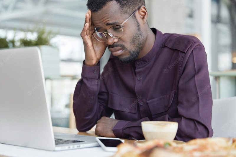 Overworked tired dark skinned male with frustrated expression, looks desperately at screen of laptop, works on business project, drinks coffee not to feel sleepy. Free Photo