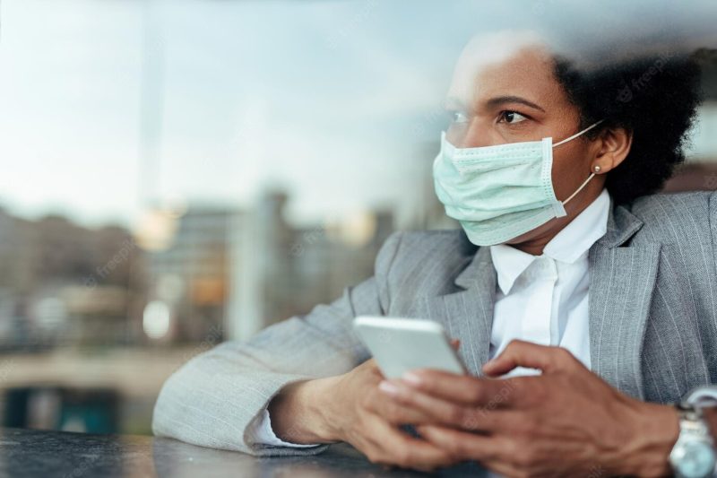 Pensive african american businesswoman with protective mask looking through the window while texting on smart phone the view is through the glass Free Photo