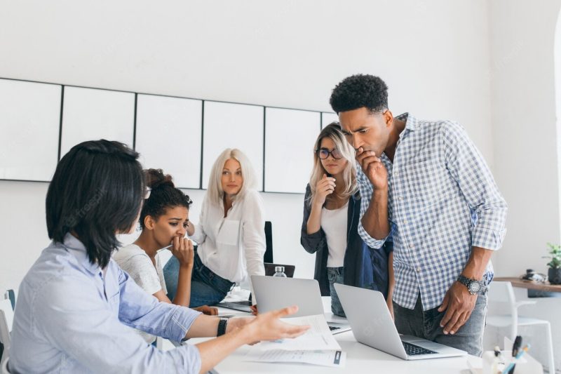 Pensive african office worker covering mouth with hand, while solving problem with computer. team of asian and black web-programmers found a mistake in their project. Free Photo