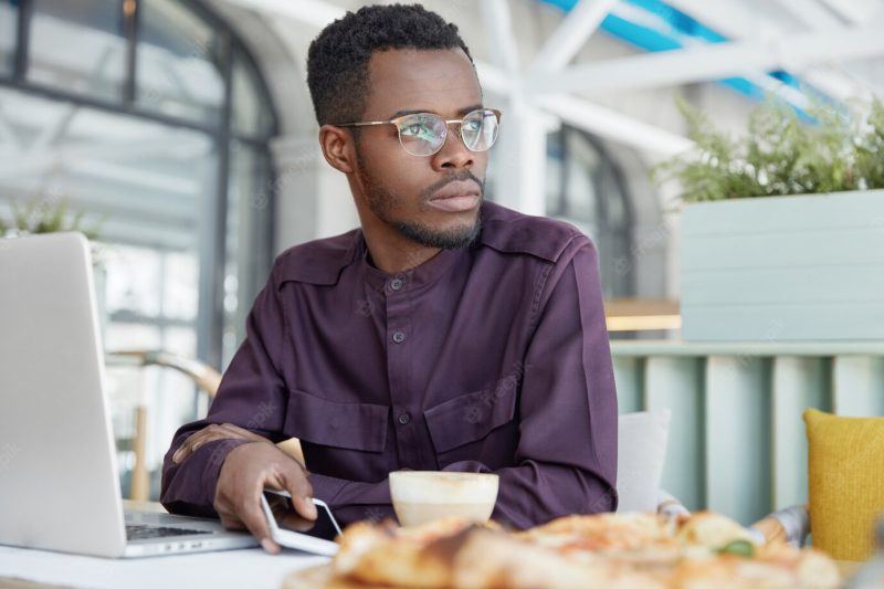 Pensive dark skinned African male freelancer in elegant shirt, works on new project, holds modern smart phone, drinks coffee Free Photo