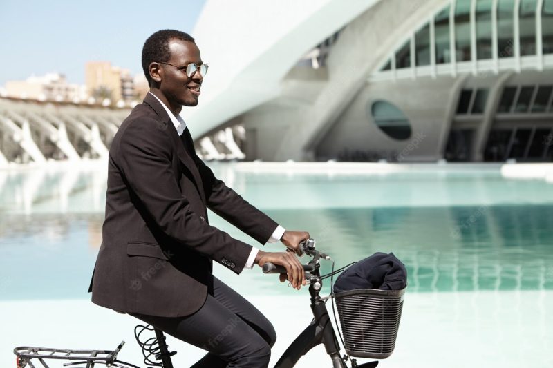 People, modern lifestyle, transport and ecology concept. successful ecologically friendly dark-skinned male head of large financial company going to office on bicycle, wearing black formal suit Free Photo