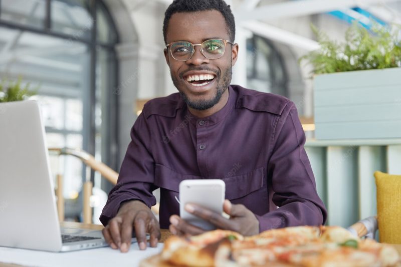 Photo of dark skinned prosperous office manager uses modern cellular for sending messages, works on portable laptop computer Free Photo