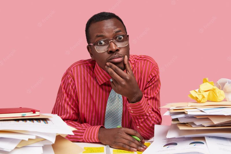 Photo of handsome dark skinned male director wears formal shirt and tie, holds chin, works on financial report at desk, has piles of documentations Free Photo