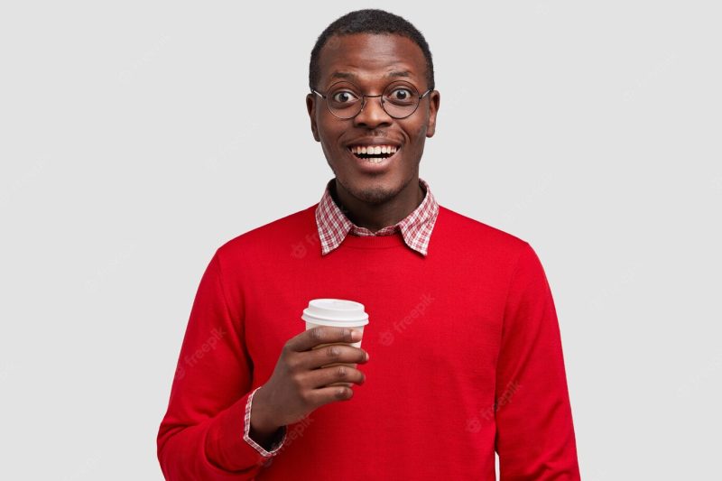 Photo of handsome smiling dark skinned young man dresssed in red jumper, holds takeaway coffee, being in good mood Free Photo