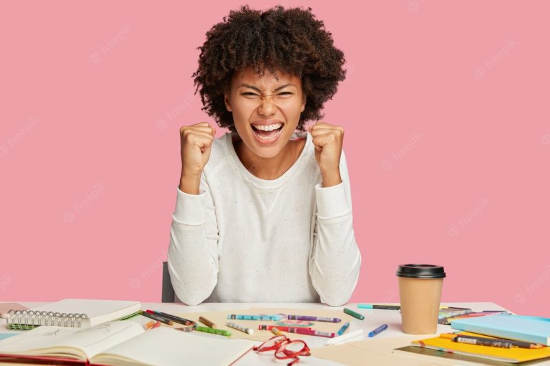 Photo of overjoyed African American architect keeps fists clenched, smiles broadly, squints face Free Photo