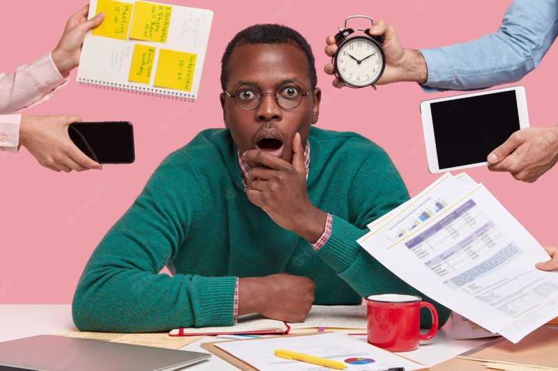 Photo of scared dark skinned guy holds chin, opens mouth widely, wears glasses, surrounded with peoples hands holding electronic gadgets Free Photo