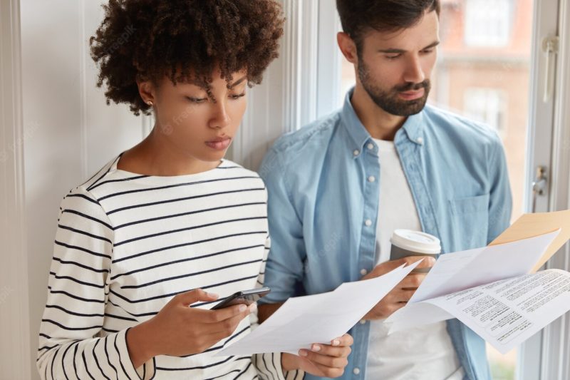 Photo of serious black woman studies checklist, uses calculator on mobile phone, unshaven guy with bristle holds coffee, learns working audit, prepares annual financial report, checks inspection Free Photo