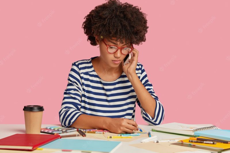 Photo of serious busy female worker with afro hairstyle, creats illustration for project work, talks with partner via cellular, wears transparent glasses and striped clothes, isolated over pink wall Free Photo