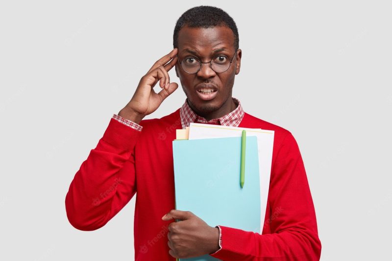 Photo of stressful displeased dark skinned young guy kepes hand on temple, frowns face, dressed in red elegant sweater Free Photo