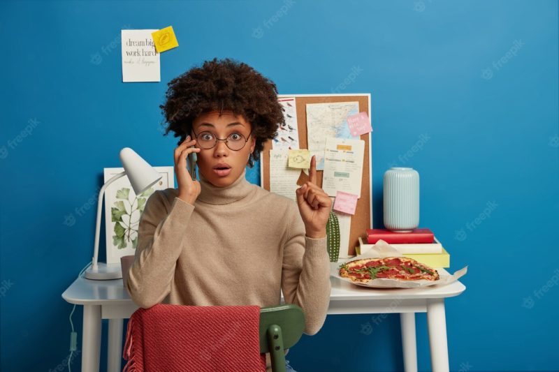 Photo of surprised shocked female worker in glasses points up during phone conversation, calls someone via smartphone, sits at chair against desktop with pizza, board with notes, desk lamp, blue wall Free Photo