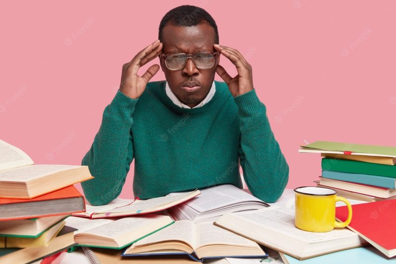 Photo of upset black young male worker has headache, works hard, reads literature, suffers from migraine Free Photo