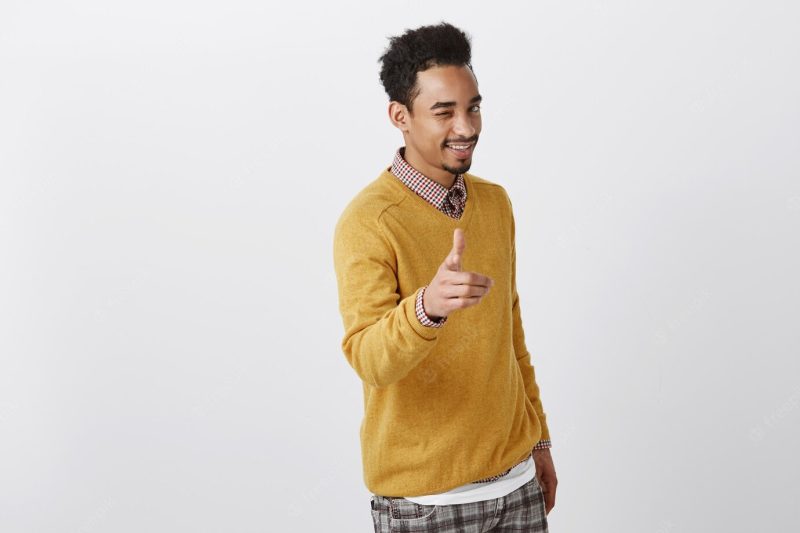 Picking you to be partner in crime. charming emotive African-American male with Afro hairstyle in yellow pullover showing gun gesture, smiling broadly, greeting woman in club Free Photo